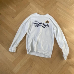 Vit stickad tröja från Morris Company - Snygg vit stickad tröja från Morris Company med broderad logga och text på bröstet. Tröjan har ribbade muddar vid ärmslut och nederkant samt rund halsringning. Perfekt för dig som gillar klassisk och stilren look.