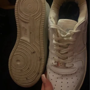 Nike Air Force 1 vita sneakers - Klassiska Nike Air Force 1 sneakers i helvitt skinn med perforerad tå och snörning. Snygg och tidlös modell med låg profil och rund tå. Perfekta för dig som vill ha en clean och ikonisk look till vardags. AF1-detalj på snörningen och Nike-logga på sidan.