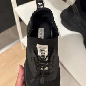 Svarta sneakers från Steve Madden - Svarta chunky sneakers från Steve Madden med stickad mesh-ovandel och grov platt sula. Skorna har snörning med elastiska snören och en vit logotypdetalj på plösen. Perfekt för dig som gillar sportig och modern stil.