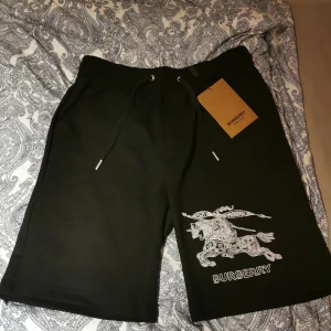 Burberry - Svarta shorts från Burberry med snyggt broderat riddarmotiv och logga i vitt på ena benet. Modellen har snörning i midjan och är tillverkad i mjuk bomull, perfekt för en chill och stilren look.  Pris kan diskuteras 
