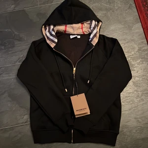Svart Burberry hoodie med dragkedja - Svart hoodie från Burberry i storlek M med klassiskt rutigt foder i huvan och guldfärgade detaljer på dragkedjan. Tröjan har snörning i huvan och ribbade muddar. Perfekt för dig som vill ha en stilren och lyxig look. Inte rakt på storlek då det känns som en S men är M