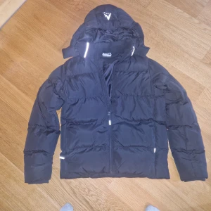 Svart pufferjacka från McKinley - Säljer en svart pufferjacka från McKinley med huva och reflekterande detaljer. Jackan har dragkedja framtill och är vadderad för extra värme. Perfekt för kalla dagar och har en clean, sportig look.