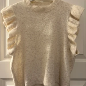 Beige stickad tshirt  Noisy May - Supersöt beige stickad tröja från Noisy May med volangdetaljer på axlarna. Ärmlös modell med ribbad halsringning och mjukt material. 