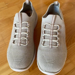 Beige sneakers från Rieker MemoSoft - Snygga beige sneakers från Rieker MemoSoft med stickad ovandel i syntetmaterial. Skorna har elastiska snören i vitt och beige, samt vit sula med grå detalj. Lätta och flexibla med sportig känsla och diskret randig detalj på häl och plös. Använd två gånger. Nypris 700-900:-