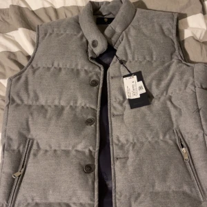 Grå dunväst från IZAC med knappar - Snygg grå dunväst från IZAC med hög krage, knäppning med både knappar och dragkedja samt två fickor med dragkedja. Västen har en stilren look och är fodrad med mönstrat tyg på insidan. Perfekt för lager-på-lager under kyliga dagar.