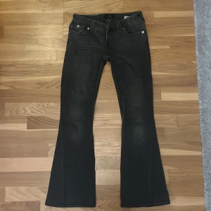 True Religion Joey Low Rise Flare Jeans - Svarta jeans från True Religion, modell Joey Low Rise Flare. Byxorna har låg midja och utsvängda ben med klassiska fem fickor och silverfärgade knappar. Snygg tvättad look och detaljerade bakfickor. Perfekta för dig som gillar retrovibbar och statement-jeans. Byxorna är korta! Jag är 160 och dom är lite korta på mig.