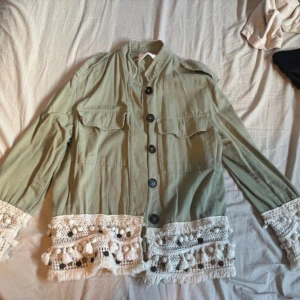 Grön utilityjacka med virkade detaljer - Snygg ljusgrön utilityjacka med stora bröstfickor och svarta knappar framtill. Jackan har unika virkade spetsdetaljer med tofsar och bollar i vitt längs nederkant och ärmslut. Rak passform och lång ärm, perfekt för dig som vill sticka ut. Den har ett litet litet märke/hål där fram men som är trådigt så inte ett helt tomt hål , inget synligt när jackan är på, annars jättefint skick!!! Kom med prisförslag !!!