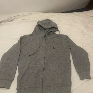 Grå hoodie  - Snygg grå hoodie från Polo Ralph Lauren med dragkedja framtill, huva med snörning och klassisk logga på bröstet. Tillverkad i mjuk bomullsblandning och har praktiska fickor, varav en med dragkedja på insidan. Perfekt för chill dagar.