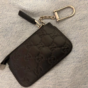 Svart Gucci plånbok i skinn - Snygg svart plånbok från Gucci i äkta skinn med präglat GG-mönster över hela ytan. Praktisk dragkedja och silverfärgad kedja med karbinhake, perfekt att fästa i väskan eller byxan. Stilren och lyxig accessoar för dig som gillar designer.