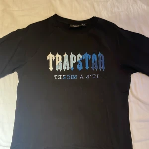 Svart Trapstar t-shirt med blått tryck - Svart t-shirt från Trapstar i storlek M med ikoniskt tryck på bröstet i blått och vitt. Klassisk rund halsringning och korta ärmar. Skön passform och tillverkad i mjukt bomullstyg. Perfekt för dig som gillar streetwear och vill sticka ut.