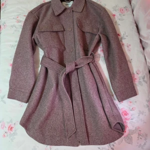 Kappa med bälte från ONLY - Säljer en brun overshirt från ONLY i storlek XS. Jackan har klassisk krage, två stora bröstfickor, knappar framtill och ett matchande bälte i midjan. Materialet är mjukt och känns ulligt, perfekt för lager-på-lager. Långärmad och med snygg loose passform.