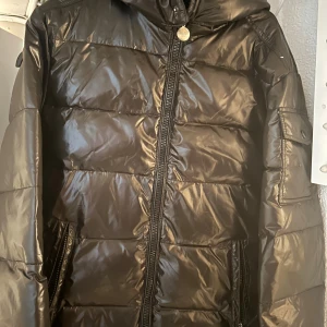 Svart pufferjacka med huva - Säljer en svart glansig pufferjacket, i fint skick men ett litet märke på armen efter jag tog bort ett annat märke. Men den är ren och fräsch.