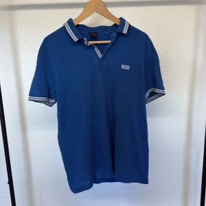 Blå pikétröja från Hugo Boss L  - Snygg blå pikétröja från Hugo Boss med vit logga på bröstet. Tröjan har klassisk krage med vita och grå ränder samt matchande detaljer vid ärmsluten. Perfekt för en stilren och sportig look.