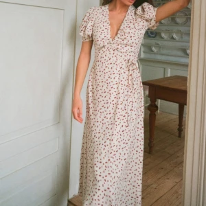  Adoore ferrone Cherry  - Söt långklänning i ljus färg med små röda körsbär, omlottmodell och knyt i midjan. Klänningen har puffiga kortärmar och djup v-ringning. Perfekt för sommarens alla tillfällen. Passar S !