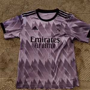 Real Madrid Adidas lila matchtröja S - Säljer en lila Real Madrid fotbollströja från Adidas i storlek S. Tröjan har korta ärmar, svart krage och svarta detaljer på axlarna. Mönstret är grafiskt med ljusa och mörka lila nyanser. Materialet är lätt och andas, perfekt för träning eller match.
