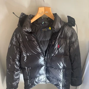 Svart pufferjacka från Polo Ralph Lauren - Svart glansig pufferjacka från Polo Ralph Lauren med huva och dragkedjor på fickorna. Jackan har en röd broderad logga på bröstet och är vadderad för extra värme. Perfekt för kalla dagar och har en modern, sportig look. Nypris är 5500 priset är ej hugget i sten. 