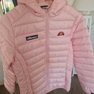 Ljusrosa pufferjacka från Ellesse - Säljer en ljusrosa pufferjacka från Ellesse med huva och dragkedja framtill. Jackan har Ellesse-logga på bröstet och är quiltad för extra värme. Perfekt för kyliga dagar och riktigt snygg färg som sticker ut.