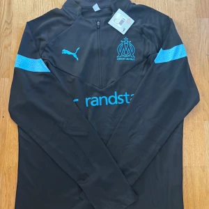 Marseille långärmad träningströja Puma - Svart långärmad fotbollströja från Puma för Olympique de Marseille med ljusblå detaljer och klubbmärke på bröstet. Halvlång dragkedja framtill och tryckt sponsorlogga. Tillverkad i lätt och funktionellt material.