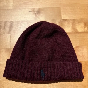 Vinröd stickad mössa från Polo Ralph Lauren - Vinröd stickad mössa från Polo Ralph Lauren med klassisk ribbad uppvikning och broderad blå logga framtill. Perfekt accessoar för kyliga dagar och enkel att matcha med olika outfits.