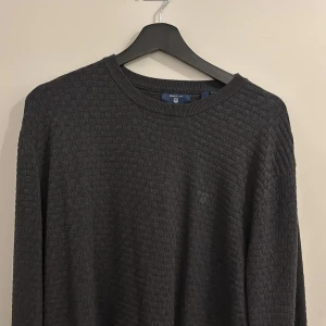 Mörkgrå stickad tröja från GANT  - Säljer en mörkgrå stickad tröja från GANT. Tröjan har ett diskret rutmönster i strukturen och rund halsringning. Perfekt för dig som gillar klassisk och stilren design med en twist. Storlek XL men passar bättre som L
