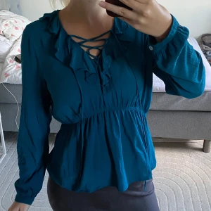 Petrolblå blus med volang och snörning - Snygg petrolblå blus med volangdetaljer vid halsringningen och trendig snörning framtill. Långärmad modell med lätt rynkad midja som ger en fin siluett. Perfekt för dig som vill ha en feminin och cool look.