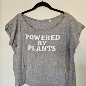 Grå t-shirt med texttryck - Grå t-shirt med vit text 'Powered by Plants' framtill. T-shirten har rund halsringning och korta ärmar. Tillverkad i mjuk bomull.