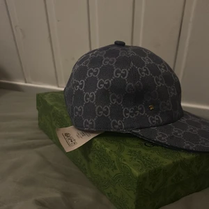 Gucci keps i blått GG-mönster - Säljer en exklusiv blå keps från Gucci med ikoniskt GG-mönster över hela kepsen. Kepsen har en böjd skärm och är tillverkad i bomullsmaterial. Liten guldfärgad GG-detalj på sidan. Kommer med original grön Gucci-box.