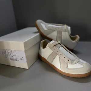 Maison Margiela sneakers i vitt och beige - Maison Margiela sneakers i vitt och beige med klassisk gummisula. Skorna har detaljer i mocka och skinn, samt en stilren design utan synliga loggor på ovandelen. Perfekta för dig som gillar minimalistisk och trendig stil.