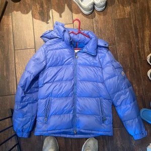 Blå Moncler pufferjacka med huva passar storlek M - Snygg blå pufferjacka från Moncler med huva och dragkedja framtill. Jackan har två fickor med dragkedja och en ärmficka med Moncler-logga. Perfekt för kalla dagar och riktigt skön att bära.