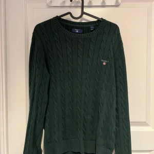 Mörkgrön kabelstickad tröja från GANT - Säljer en mörkgrön kabelstickad tröja från GANT med rund halsringning och diskret logga på bröstet. Tröjan har lång ärm och ribbade muddar, perfekt för dig som gillar klassisk och stilren look.