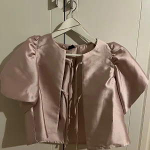 Rosa bolerotopp i satin - Supersnygg bolerotopp i rosa satin med puffiga korta ärmar och knytband framtill. Perfekt för dig som vill sticka ut med en elegant och trendig look. Toppens glansiga material ger en lyxig känsla och den croppade modellen är riktigt modern. Helt ny, använd bara 1 gång så väldigt bra skick. Pris 150kr 
