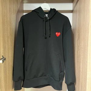 Svart hoodie Comme des Garçons Play - Svart hoodie från Comme des Garçons Play med klassiskt rött hjärta med ögon broderat på bröstet. Tröjan har huva med snörning. Pris satt ex. frakt