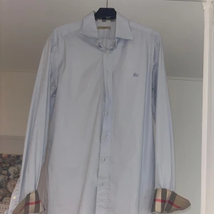 Burberry Skjorta - Tja, säljer denna Burberry skjorta i färgen ljusblå & i storlek M. Sparsamt använd, finns en mindre defekt på höger sida arm (syns på sista bilden). Mitt pris ligger på 599kr, priset går att diskutera.