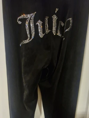 Svarta velourbyxor Juicy Couture XS - Svarta velourbyxor från Juicy Couture i storlek XS. Byxorna har smal passform och pryds av glittrande strasstenar som bildar texten 'Juicy' på baksidan. Perfekta för dig som gillar en ikonisk och glamorös look.