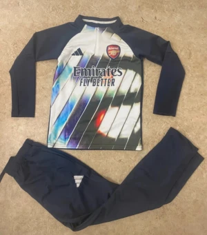 Arsenal x Adidas träningsset - Snyggt Arsenal träningsset från Adidas med långärmad tröja och matchande byxor. Tröjan har coolt mönster i blått, vitt och rött med klubbmärke och Adidas-logga, samt half zip framtill. Byxorna är mörkblå med Adidas-detalj. Perfekt för fotbollsträning.