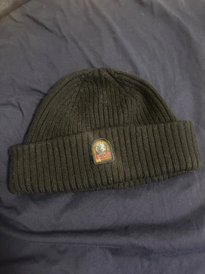 Svart ribbad mössa från Parajumpers - Svart ribbstickad mössa från Parajumpers med uppvikt kant och snyggt märke framtill. Mössan har en klassisk form och är tillverkad i mjuk akryl, perfekt för kalla dagar. Röd etikett med Parajumpers-logga på insidan ger extra stilpoäng.