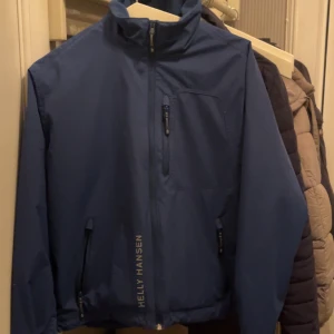 Helly Hansen  - Snygg marinblå vindjacka från Helly Hansen med dragkedja framtill, två sidofickor med dragkedja och logga på bröstet och längs sidan. Jackan är tillverkad i polyester och har en klassisk, sportig look med lång ärm och normal passform. Helt ny nästan 