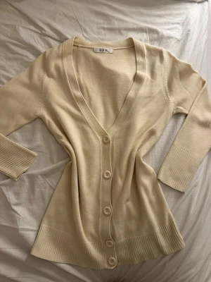 Beige kofta från SPB med v-ringning - Snygg beige kofta från SPB med djup v-ringning och stora matchande knappar framtill. Långärmad modell med breda ribbade muddar vid ärmslut och nederkant. Mjuk och tunn stickad kvalitet som känns chill och enkel att styla.