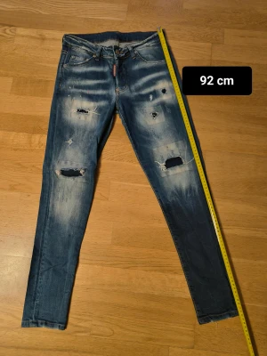 Dsquared2 blå skinny jeans med slitningar - Snygga blå jeans från Dsquared2 med skinny passform och låg midja. Jeansen har slitna detaljer, hål och färgstänk på både fram- och baksida, samt klassisk femficksdesign. Materialet är jeans med en tvättad look och Dsquared2-logga på bakfickan.