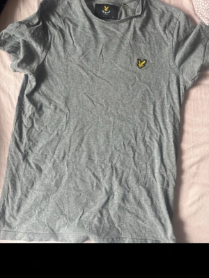 Grå t-shirt från Lyle & Scott - Säljer en stilren grå t-shirt från Lyle & Scott med klassisk gul logga på bröstet. T-shirten har rund hals och korta ärmar, perfekt för en chill och enkel look. Materialet är mjuk bomull som känns skönt mot huden.
