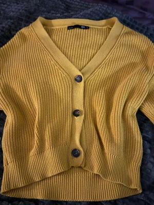 Gul croppad kofta från 157 - Säljer en snygg gul croppad kofta från 157 i storlek M. Koftan är stickad med ribbad struktur, har v-ringning och tre svarta knappar framtill. Långärmad och perfekt för att ge lite färg till din outfit. Passar dig som gillar trendiga och bekväma plagg.