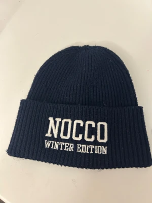 Mörkblå NOCCO mössa med broderi - Snygg mörkblå ribbstickad mössa från NOCCO med vit broderad text 'WINTER EDITION' framtill.  Från förra året julkalender därav väldigt limiteradKlassisk form med uppvikt kant, perfekt för kalla dagar. Stilren och enkel design som passar till de flesta jackor och outfits.