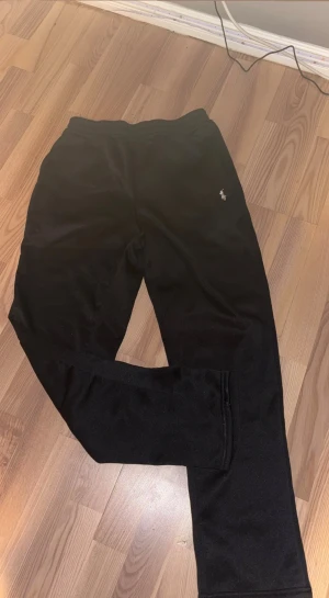 Svarta mjukisbyxor från Polo Ralph Lauren - Svarta mjukisbyxor från Polo Ralph Lauren med klassisk broderad logga i vitt på höften. Byxorna har elastisk midja och är gjorda i mjukt polyester-material som är skönt mot huden. Perfekta för chill eller träning.