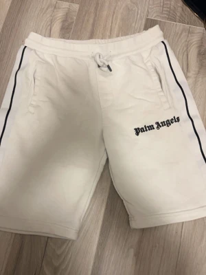 Vita Palm Angels shorts med svarta detaljer - Säljer ett par vita shorts från Palm Angels med svarta ränder längs sidorna och logga på ena benet. De har snörning i midjan och två fickor framtill. Perfekta för en avslappnad och trendig look.