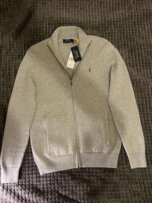 Grå ribbad kofta från Polo Ralph Lauren - Snygg grå kofta från Polo Ralph Lauren med ribbad struktur och diskret logga på bröstet. Tröjan har hel dragkedja, ståkrage och två fickor fram. Perfekt för dig som gillar stilrena och klassiska plagg med premiumkänsla.