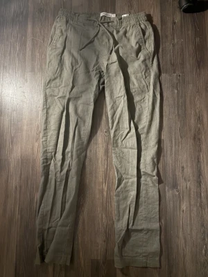 Olivgröna linnebyxor med snörning - Snygga olivgröna linnebyxor från Dressman i storlek S. Byxorna har elastisk midja med snörning och sidofickor, vilket ger en chill och avslappnad vibe. Perfekta för dig som gillar en enkel och stilren look med naturligt material.