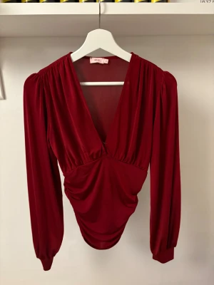 Vinröd omlottopp från Nelly - Säljer en vinröd omlottopp från Nelly med djup v-ringning och långa puffärmar. Toppen har snygg rynkning i midjan och är tillverkad i mjuk polyester med lite glans. Perfekt för dig som gillar statement-plagg med cool siluett och elegant touch. Såg att på sista vilden så ser tröjan jätte genomskinlig ut men det är inget man märker av i verkligheten. Säljer den då den aldrig används😊