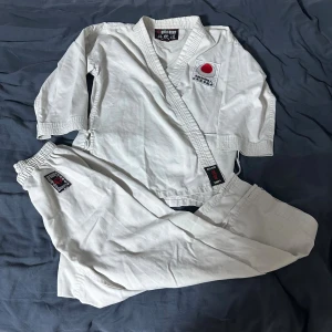 Vit Budo-Nord karategi storlek 150 - Säljer en klassisk vit karategi från Budo-Nord i storlek 150. Dräkten har broderade japanska tecken och röd logga på både jacka och byxa. Tillverkad i slitstark bomull med förstärkta sömmar för extra hållbarhet. Perfekt för träning och tävling.
