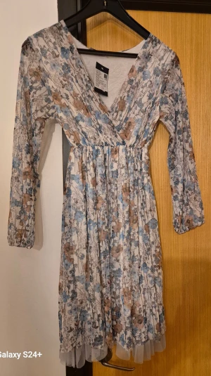 Blommig kortklänning från Lana Griss - Supersöt kortklänning från Lana Griss med blommigt mönster i blått, beige och brunt. Klänningen har omlott-v-ringning, långa ärmar och ett skirt lager längst ner. Perfekt för dig som gillar romantisk och boho-inspirerad stil.