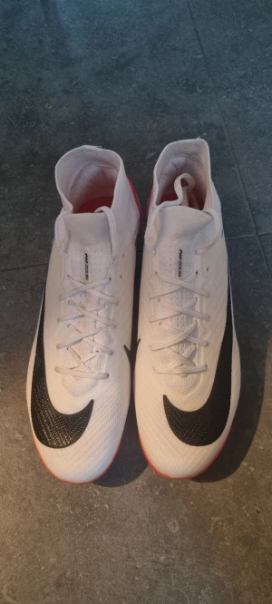 Nike Air Zoom fotbollsskor vit/röd - Nike Air Zoom fotbollsskor i storlek 42, med en clean vit ovandel och röda detaljer på sulan. Svart Nike Swoosh på sidan och snörning framtill. Skorna har platt sula med dobbar för gräsplan och är tillverkade i syntetmaterial för lätt känsla och bra grepp.